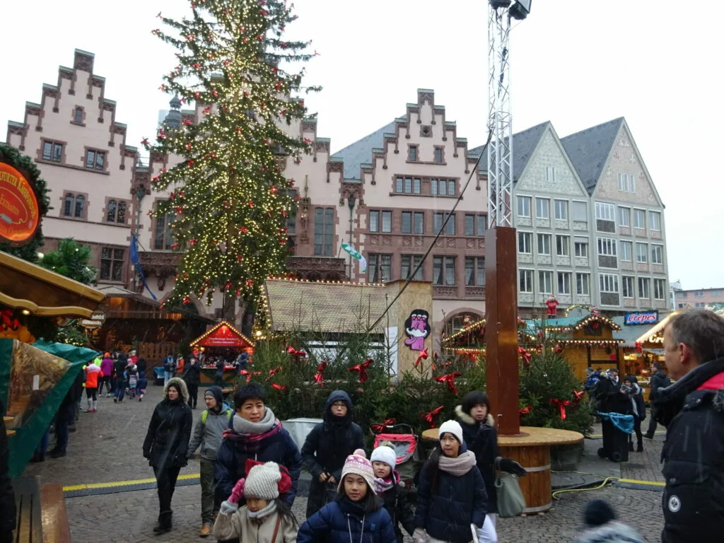 Festive Christmas market in historic town square with decorated wooden stalls, ivy-covered tree, and traditional architecture