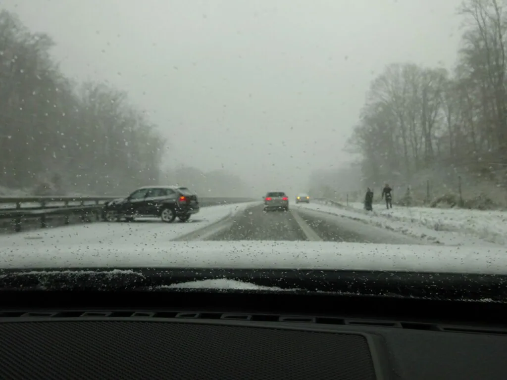 View from inside a vehicle on a snowy highway with an accident scene visible ahead, bare trees lining both sides of the road