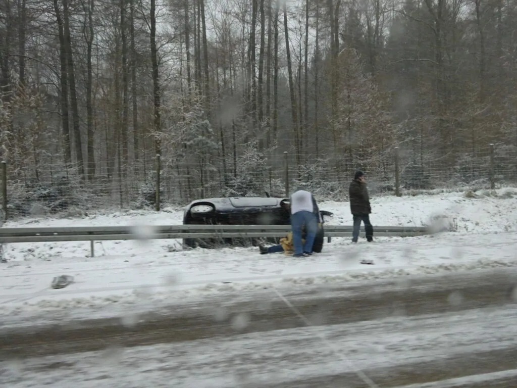 Overturned vehicle on snowy roadside with bare trees and person standing nearby