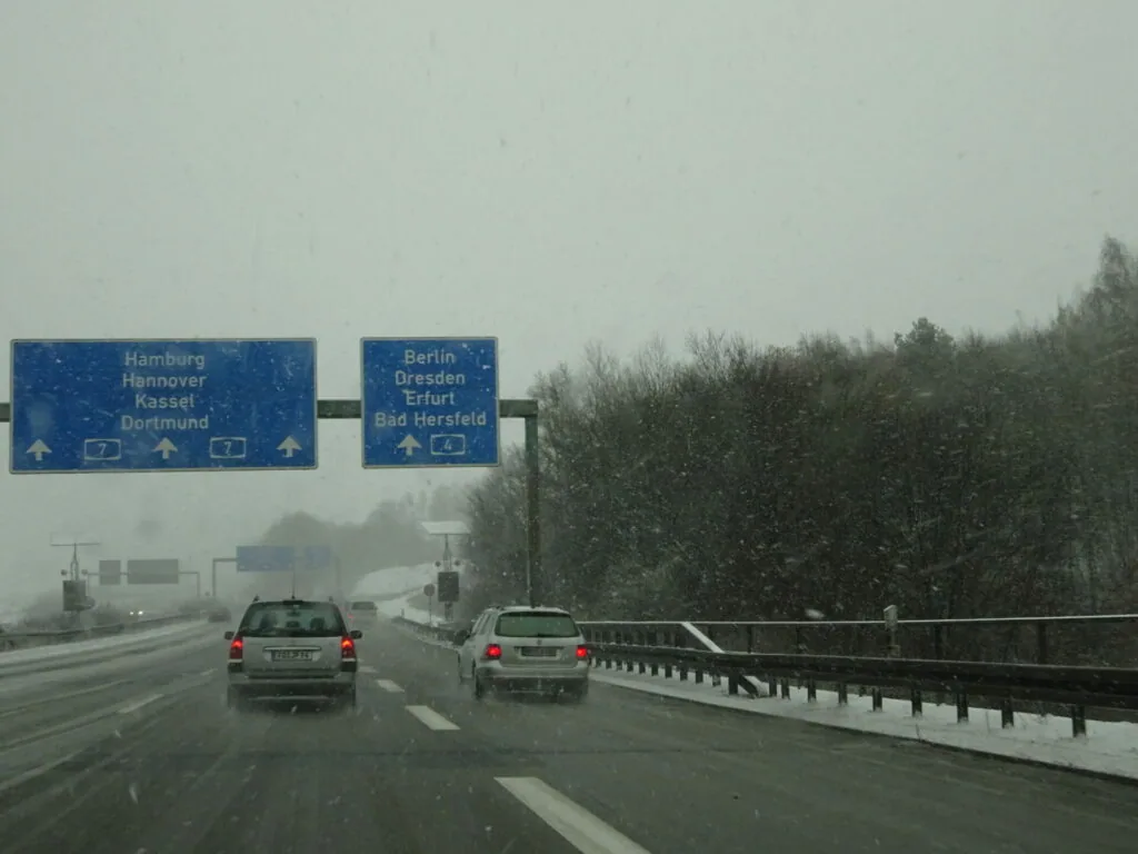 Heavy snow falling on German highway with blue directional signs for Hamburg, Berlin, and Dresden above two cars driving ahead