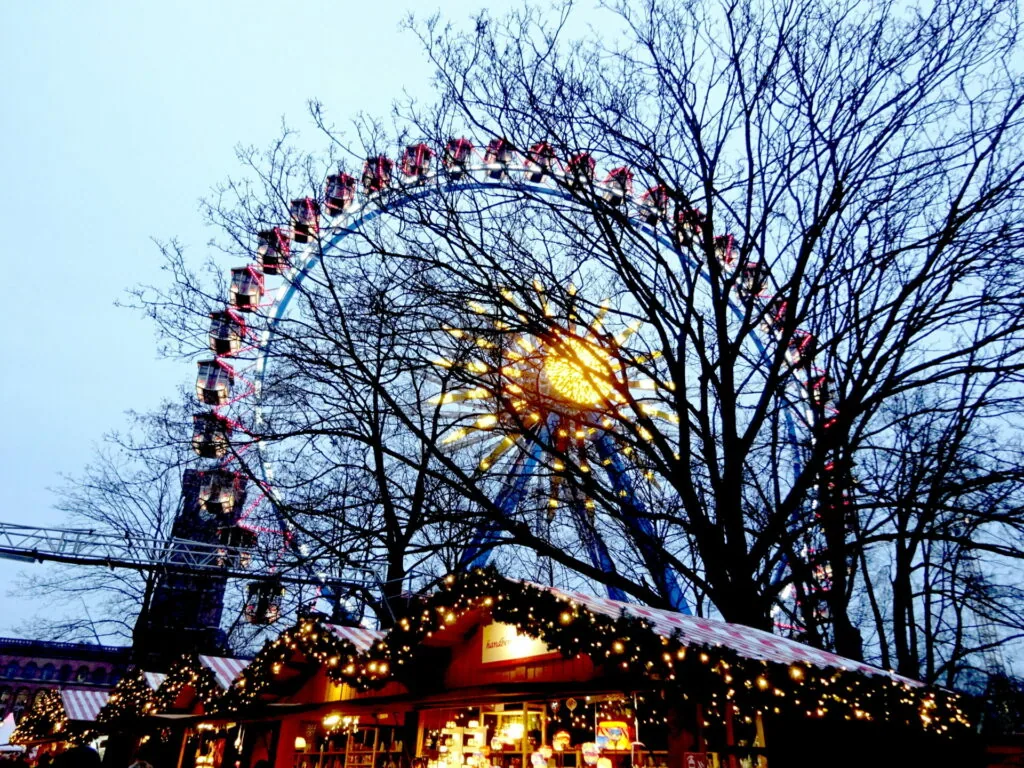 Illuminated Ferris wheel and lit wooden market structure at dusk with bare winter trees