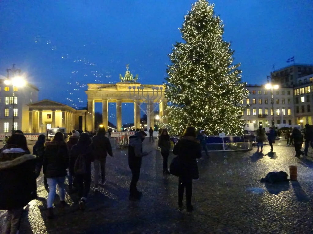 Illuminated Christmas tree in plaza at dusk with Brandenburg Gate and historic buildings lit in background, people gathered in winter scene