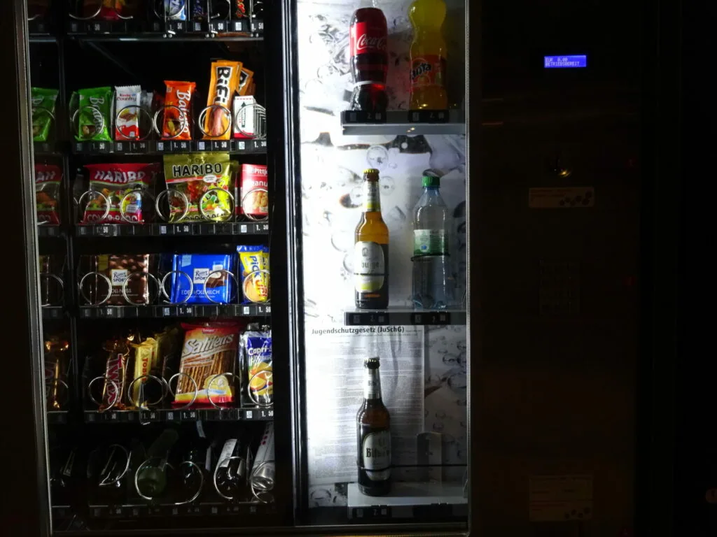 Two illuminated vending machines at night, one stocked with snacks and candy on the left, one with beverages including beer and water bottles on the right.