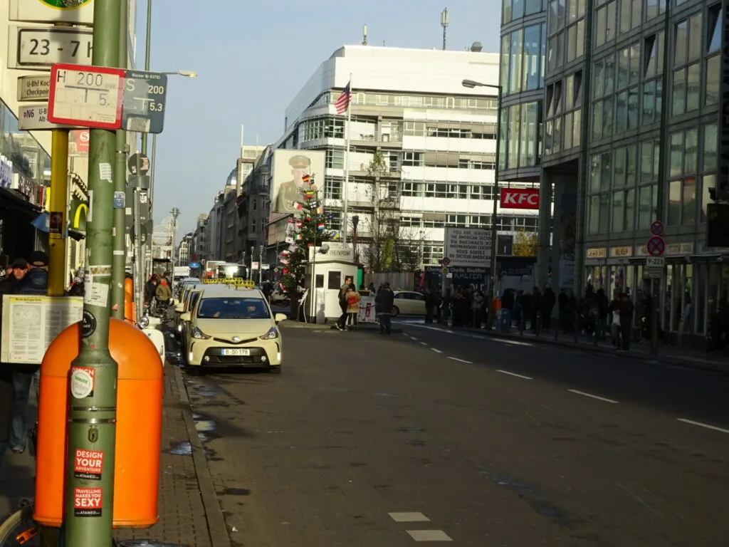 Urban street scene with white car, pedestrians, storefronts including KFC sign, and modern buildings on clear day