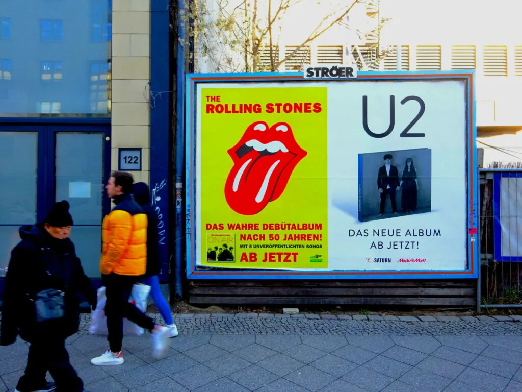 Large outdoor advertisement billboard featuring Rolling Stones and U2 album promotions with pedestrians walking past