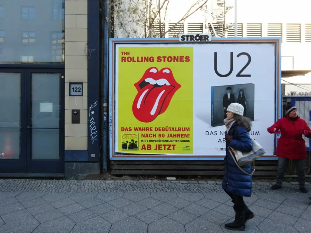 Street billboard displaying The Rolling Stones and U2 album advertisements with pedestrians walking by in winter clothing