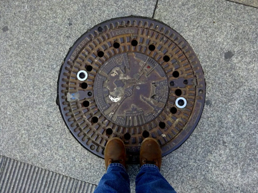 Person in brown boots and blue jeans standing on round metal manhole cover with ventilation holes