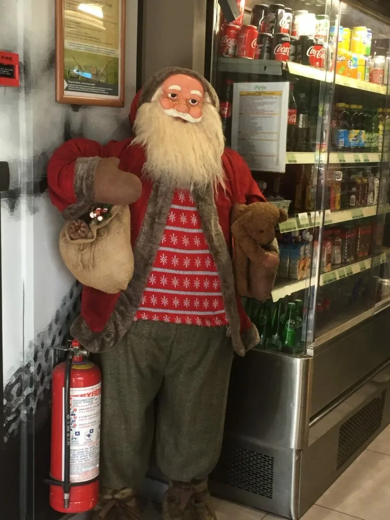 Santa Claus figure wearing red coat and patterned sweater, standing next to refrigerator display in store