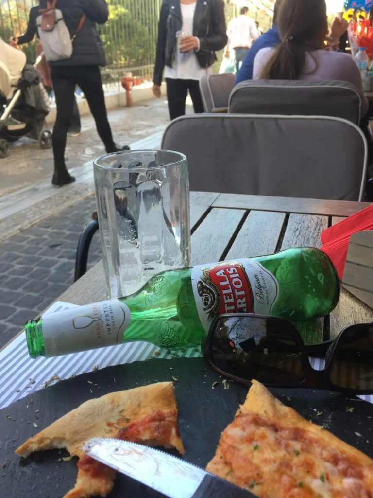 Outdoor dining table with pizza slices, beer bottles, and a glass of water, with people sitting in the background