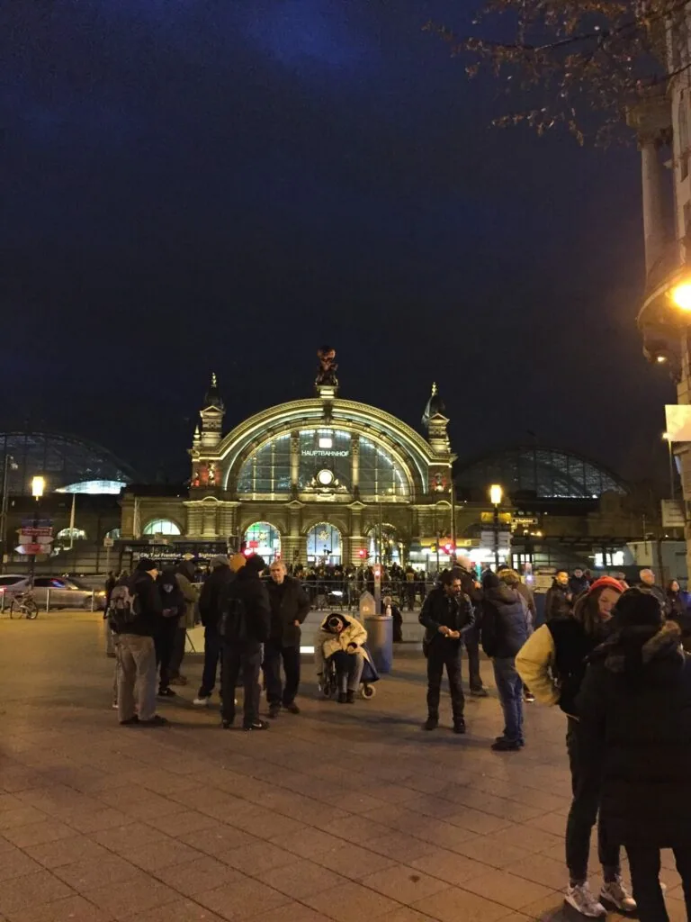 Illuminated train station facade at night with crowds of people in the plaza, ornate architecture with arched entrance and illuminated clock tower