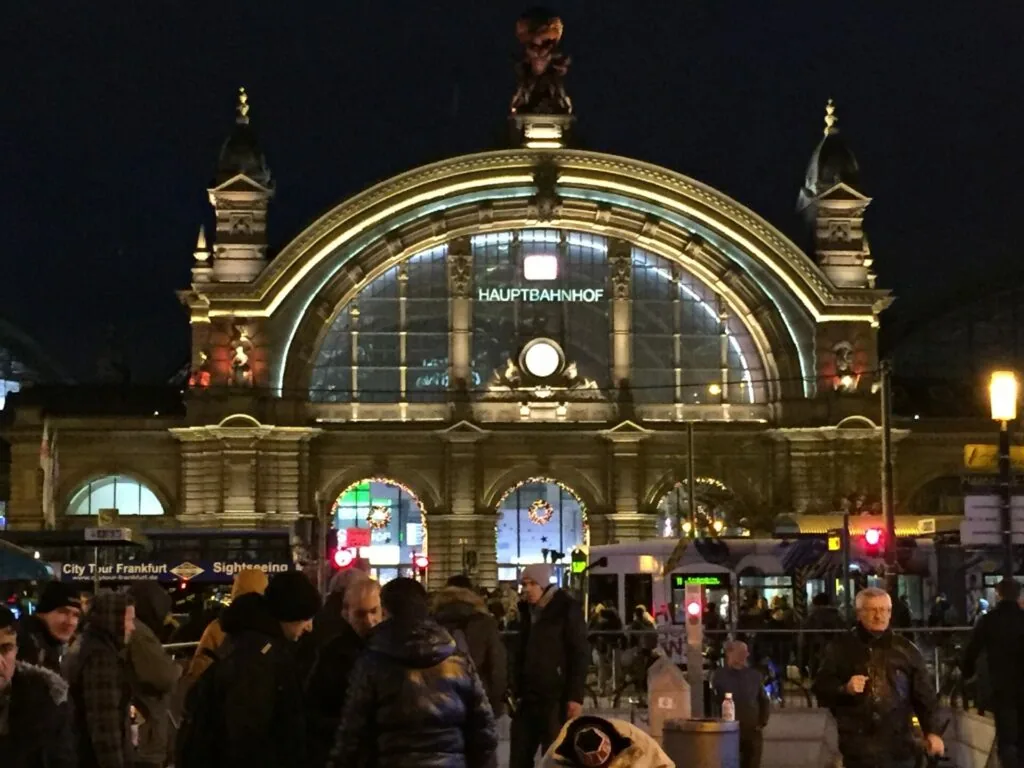 Illuminated historic railway station building with arched glass entrance at night, crowds of people in foreground
