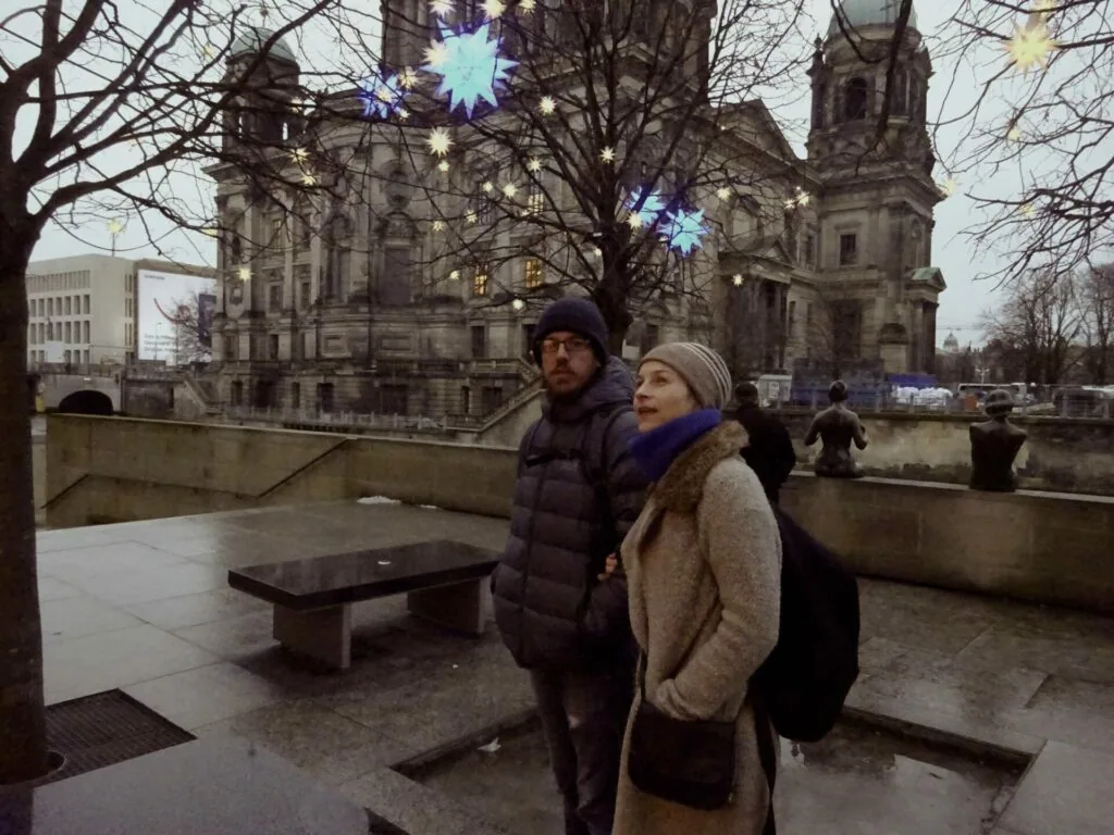 Two people in winter clothing stand on a paved plaza near bare trees, with a cathedral and decorative light installations visible in the background