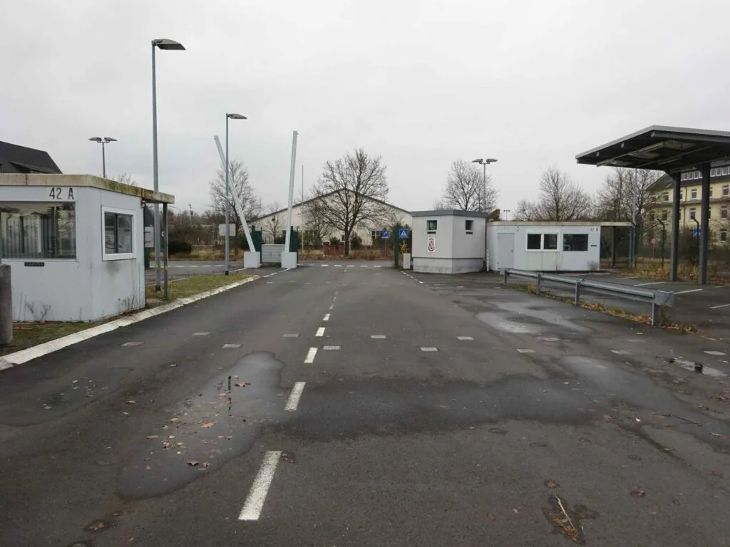 Empty asphalt road with white dashed lines, small white administrative buildings on sides, bare trees, street lights, and puddles on wet pavement