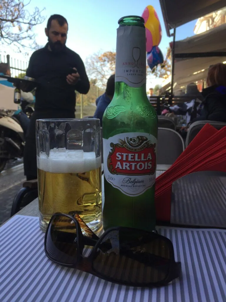 Stella Artois beer bottle and glass with foam on outdoor table, person in background